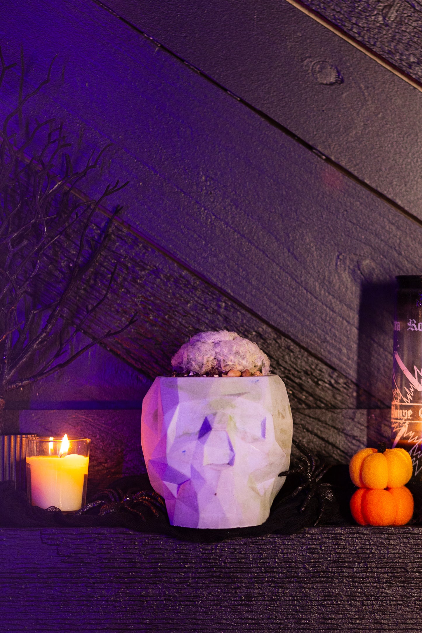 A single large cement skull planter with a geometric design sits on a dark wooden mantel. The planter is light green in color and holds a fuzzy cactus on top, giving it a spooky yet modern appearance. To the left of the planter, a yellow candle emits a warm glow, adding to the eerie ambiance. On the right, small stacked orange pumpkins enhance the Halloween theme. The background features dark wood paneling, further emphasizing the mysterious and gothic atmosphere.