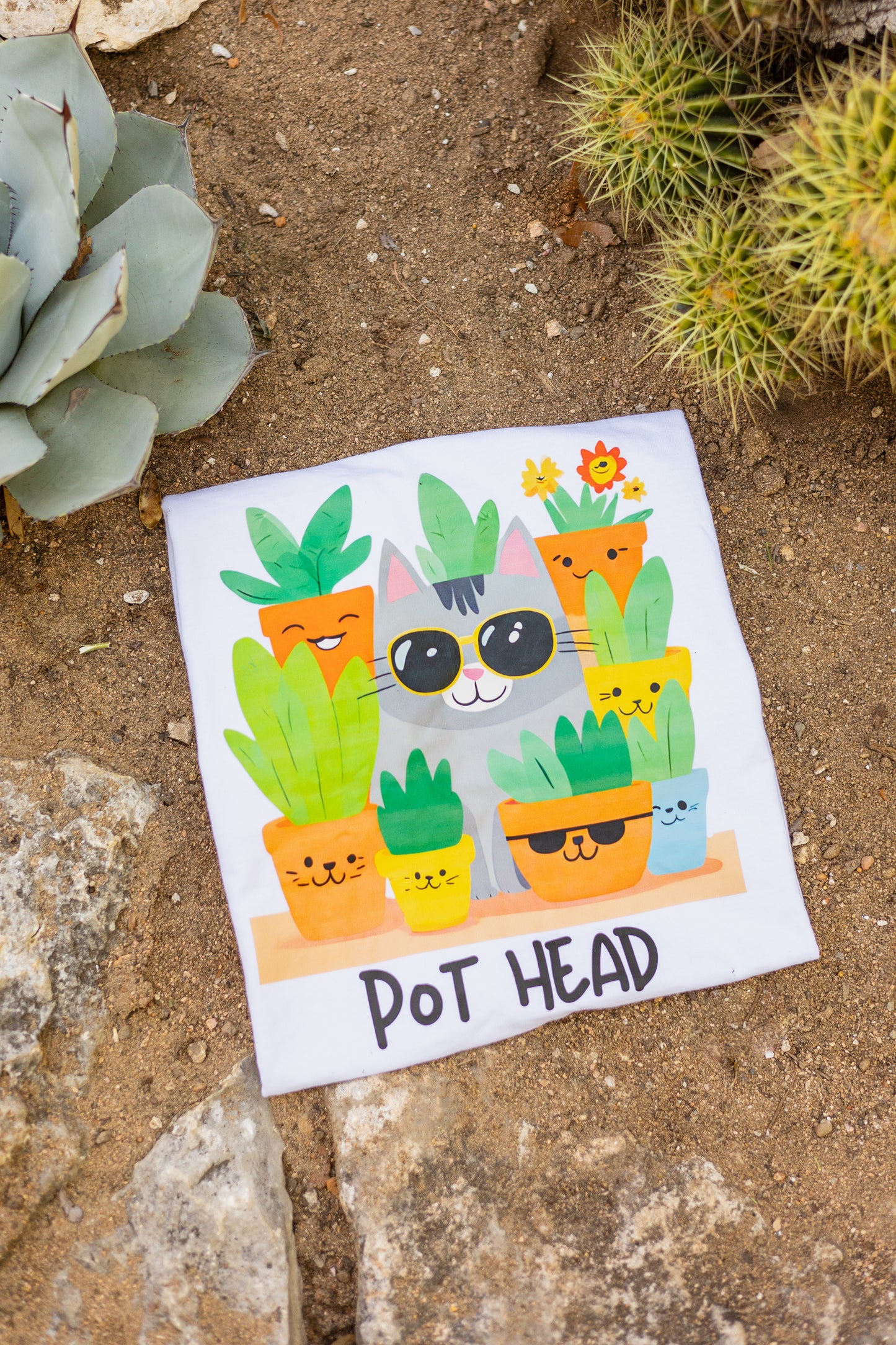 White Pot Head T-shirt flatlay with a quirky cat wearing sunglasses surrounded by plant pots, placed on a desert background surrounded by agave plants, spikey cactus, and rocks.