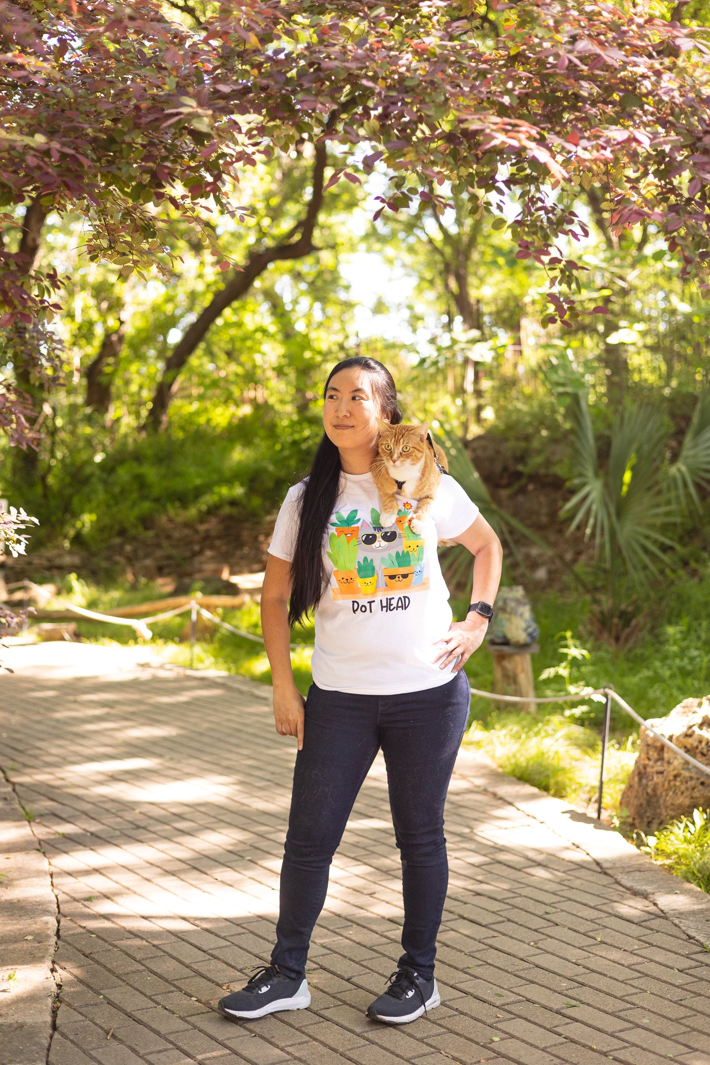Asian lady with long hair, a ginger cat on her shoulder, posing while wearing a white Pot Head T-shirt on a brick path with palms and a red maple tree in the background.