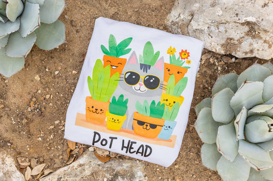 White Pot Head T-shirt flatlay with a quirky cat wearing sunglasses surrounded by plant pots, placed on a desert background surrounded by agave plants and rocks.
